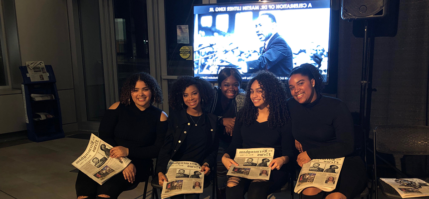 5名学生坐在一排，手里拿着一张印有马丁·路德·金照片的报纸. for a Black History Month performance.