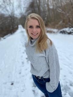 Student who identifies as female, standing in the snow, posing and smiling wearing a grey sweater with jeans. 